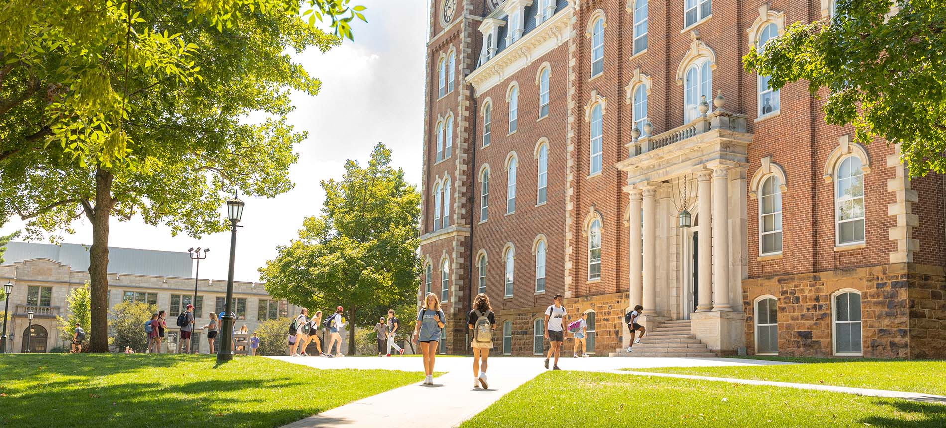 Students stroll across a sunny green lawn toward Old Main’s grand brick facade, framed by leafy trees and bright skies.