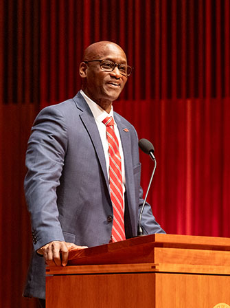 A poised university chancellor Robinson stands and speaks at a wooden podium, ready to inspire, framed by bold red curtains.