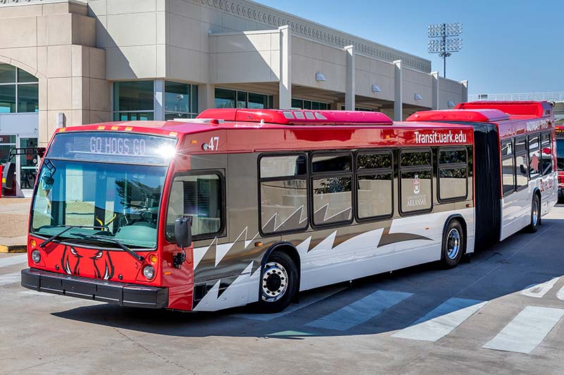Razorback Transit bus with red roof and silver body parked near a beige campus building.
