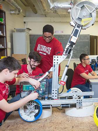 Team members build a complex robot featuring a tall arm and circular mechanism on a wheeled base in a classroom workshop.