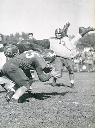 1940's photo of dramatic football tackle that sends a player soaring headfirst through the air as defenders clash fiercely on a sunlit field.