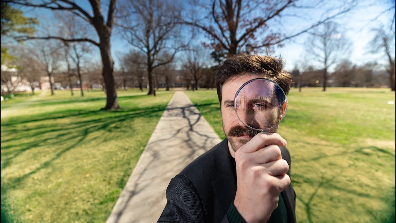 Person holding a magnifying glass on a walkway in a park with green grass and leafless trees.