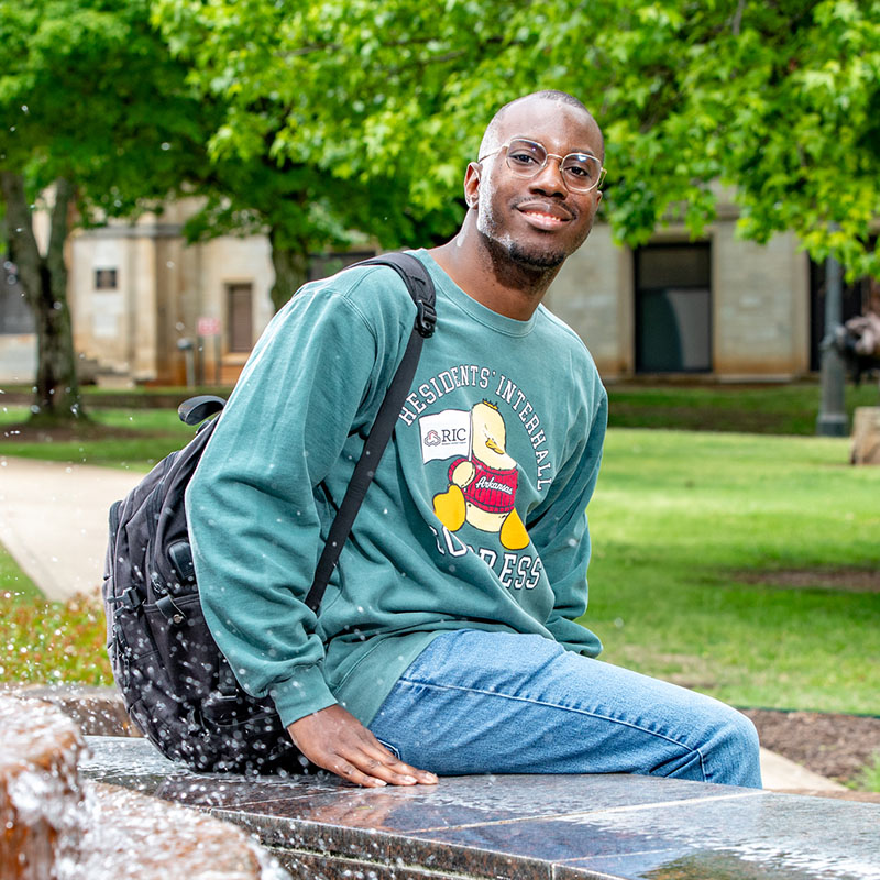Danterrion sitting by a campus fountain wearing a green sweatshirt and jeans with a backpack nearby.