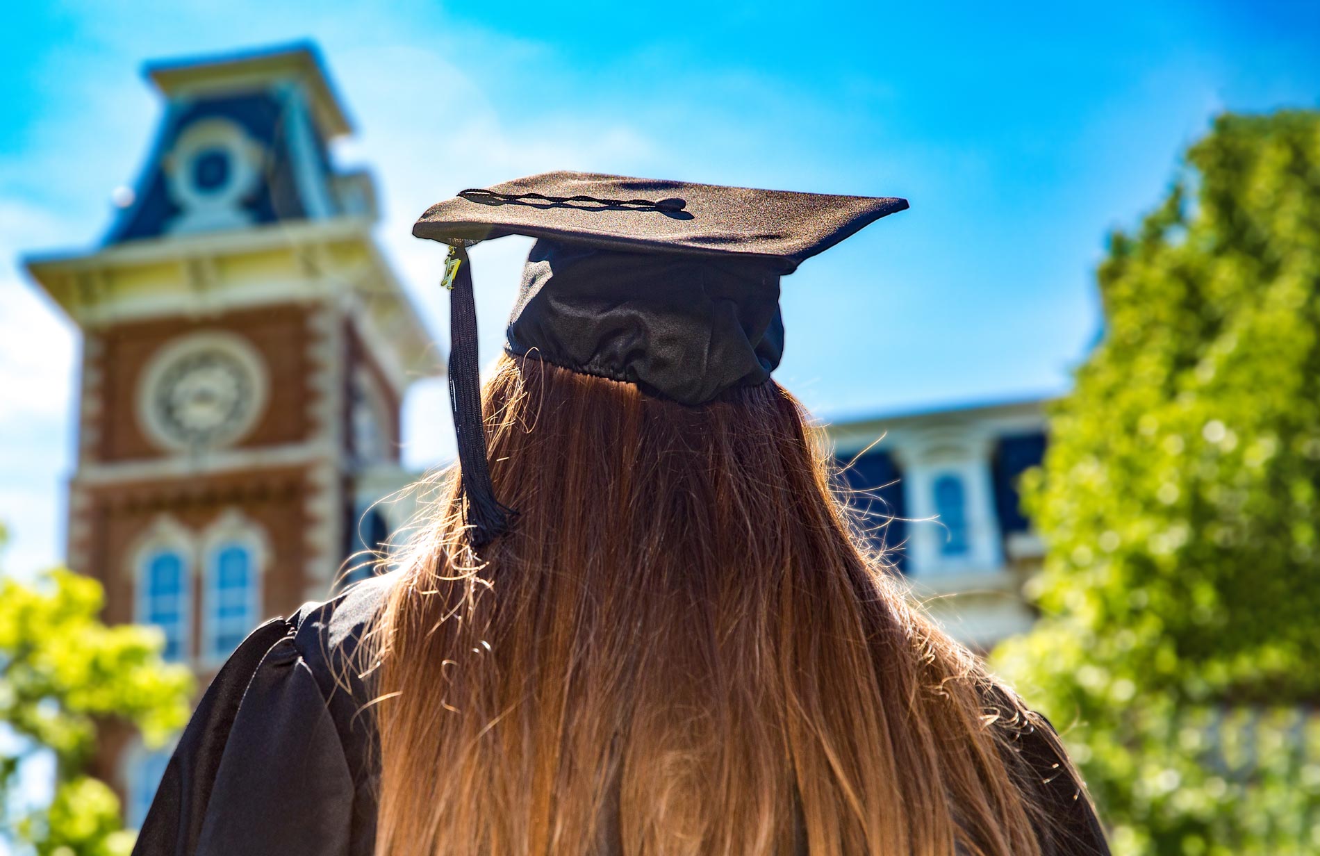Graduate in cap and gown standing outdoors with Old Main building in the background under a bright blue sky.