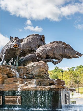 Bronze sculpture of three wild hogs on a rocky fountain with cascading water, set outdoors under a blue sky with clouds.