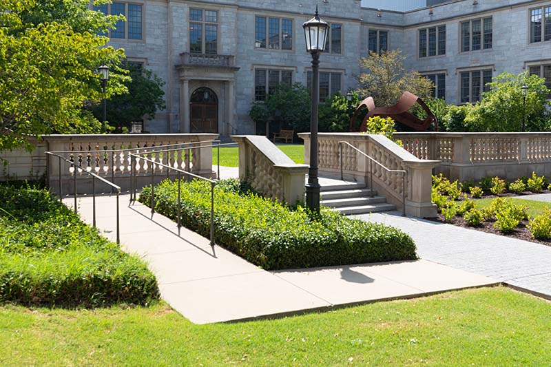 Sunlight bathes a stately university campus courtyard with stone railings, lush greenery, and a graceful ramp leading to grand steps.